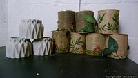 Three white geometric concrete plant pots stacked in a triangle formation with eight woodland plant pots wrapped in burlap, decorated with green leaf patterns and small red mushroom ornaments, arranged behind them.