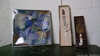 Photo showing sealed decorative plate with purple flower artwork, cheese knife in box with ribbon, and serving spatula packaged alongside.