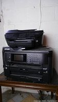 Stacked view of two black Epson printers, the top one being a multifunction scanner printer, bottom one with numeric keypad and multiple buttons.