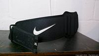 Nike XL black powerlifting belt with white swoosh logo on exterior, displayed flat on a surface showing front and size tag.