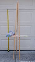 Wooden three-legged easel standing upright against a garage door, with measuring tape beside it for scale, and a transparent plastic container with small paint bottles attached at mid-level support bar.