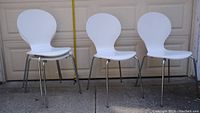 Three white dining chairs with molded seats and chrome legs seen outside against a garage door background. Chairs are modern in style with rounded backs.