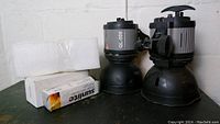 Two black and gray vintage photography lights with adjustment knobs and mounting brackets, paired with a box of Switke quartz halogen bulbs and foam protective packaging on a black surface.