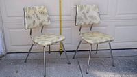 Two vintage chairs with beige tubular metal frames and padded seats and backs featuring a cream vinyl upholstery with fern pattern and glitter finish, standing outside in front of a garage door.