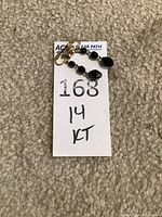 Pair of 14 kt gold clip-on earrings with black carved stones attached to a white paper labeled with lot details.