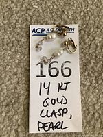 Photo of two clip-on earrings on a white card labeled '166' with handwritten note indicating '14 KT GOLD CLASP, PEARL'. Earrings feature 14 kt gold clasps, pearls, and small carved stones.