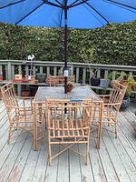 Full view of the patio furniture set including glass-top table, four rattan-style chairs, and blue umbrella on a wooden deck with plants in background.