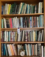 Full bookshelf with three shelves filled with assorted books and a small alarm clock on bottom shelf
