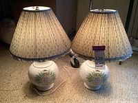 Photo showing both ceramic lamps side by side, both with round bases decorated with floral designs and French text, fabric lamp shades with blue floral pattern, left shade has missing fabric portion.