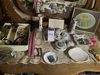 Wide view of decorative china pieces on lace cloth alongside framed floral needlepoint and assorted jewelry and bottle openers