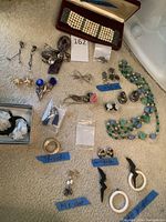Photo of entire assortment of jewelry laid out on carpet, highlighting designer labels with blue tape tags and various clip-on earrings and bracelets.