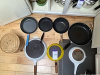 Top-down shot of multiple frying pans including blue, black, yellow, white frying pans, a bamboo steamer, and a large black skillet on wooden floor.