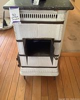 Front view of antique ceramic fireplace showing metal firebox door open, ribbed white ceramic body with dark trim, and black marble top.