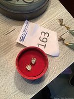 14 karat gold heart-shaped earrings with small clear stones in a red circular box on a wooden surface.