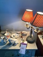 Full view of lamp with metal base and dual orange plaid fabric shades with all decorative items on tabletop including Spode china cup and saucer, ceramic puppy figurine, egg holder dish, cross and glass decanter.