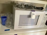 Closed white Vissani microwave oven with clear and blue glassware on the left and metal bar tools placed on top.