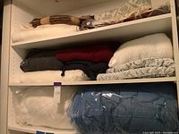 Shelf with multiple folded blankets including gray, cream, white, and a blue quilted blanket wrapped in plastic.