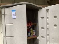 Beige Rubbermaid plastic outdoor storage shed with one door open, showing some garden chemicals and supplies on wooden shelves