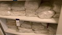 Shelves with multiple stacks of folded, white linens packaged in plastic, appearing to be bed sheets and pillowcases.