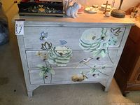 Frontal view of the small gray painted dresser with white and green flowers and small birds on the drawers and frame.
