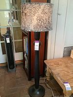 Full view of the standing lamp, showing the floral-patterned fabric shade, dark wood base, and placement beside wooden furniture.