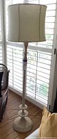 Full view of the shabby chic standing wooden lamp showing the tall distressed wood base and beige fabric shade near a window with white plantation shutters.