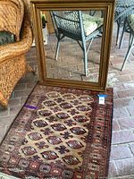Photo showing the large gilded wooden frame standing upright on a brick floor next to wicker furniture, with the Persian rug partially visible inside and underneath the frame