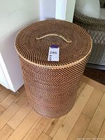 Photo of a tall cylindrical woven basket with lid and handle on hardwood floor next to wall and mirror.