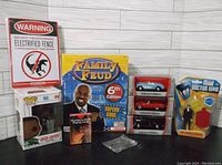 Wide shot showing multiple new in box toys including Family Feud 6th Edition game box, Green Lantern Pop Vinyl figure in box, three die-cast cars in display cases, an electrified fence warning sign, and a small boxed set.