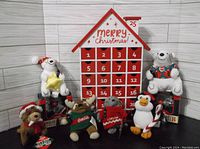 Photo of the entire lot showing two boxed Coca Cola polar bear ornaments, six plush toys including other Coca Cola polar bears and a penguin, and the red and white wooden advent calendar house with 25 numbered drawers.