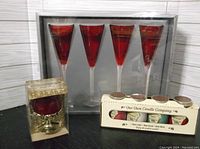 Four red stemmed glasses in original box alongside a boxed red candle in decorative metal stand, and set of four tealight candles in holder.