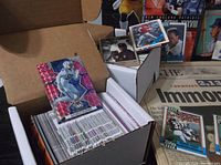 Open box containing a large quantity of primarily football trading cards, packed tightly, with one shiny red foil card displayed on top and loose cards atop the box.