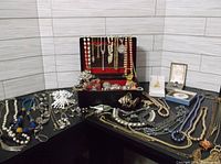Wide view of jewelry box filled with necklaces, brooches, rings, and earrings arranged on a black surface against light tiled background.