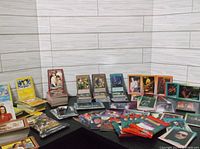 Wide shot showing the assortment of Rock n Roll, Eight Men Out, Yu Gi Oh, Star Wars, Coca Cola, and Pokemon cards laid out on a black surface against white tiled wall.