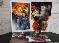 Three 11x17 posters featuring Superman, Batman, Harley Quinn and the Joker displayed together on a floor with a vinyl plank background. The posters are brightly colored and show comic book style artwork.