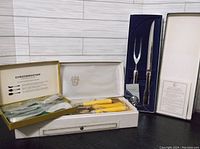Vintage carving set with steak fork and knife in box, accompanied by Towle Silversmiths cheese spreaders and Cheesemaster England cheese spreaders in original packaging.