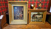 Two framed paintings displayed against patterned background, one showing an indoor scene with multiple people and the other showing two horses drinking from a water source.