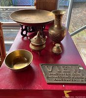 Full view of the brass vessel set on red table with window lighting, showing large plate on stand, tall vase, incense burner, small container, and one serving bowl plus another bowl partly visible.