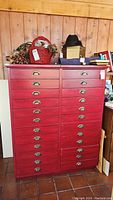 Front view of red painted wooden cabinet with two columns of fourteen drawers, each drawer fitted with a brass cup pull