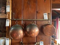 Three hammered copper pans hanging side by side on a wooden wall panel, suspended from a horizontal metal rack with ring hooks.