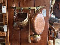 Copper cookware and wooden spoon hanging on a brass rack mounted on a wood wall, including colander and pan showing shape and patina