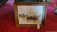 Full view of framed watercolor painting showing maritime harbor scene with boat and industrial background on red carpet.