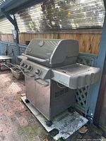 Weber Genesis propane grill with cabinet, showing metal side shelves and control knobs, located outdoors on a brick floor.