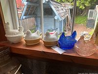 Five hen-on-nest glass bowls displayed on a wooden windowsill with a view outside.