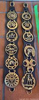 Full view of two black leather straps each holding seven brass horse brasses with different traditional motifs arranged vertically.