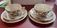 Two Limoges teacups with matching saucers and small plates decorated with floral patterns and gold trim, shown from a side view.