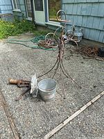 Full lot view showing rusty metal plant stand, vintage hand water pump, metal bucket, and metal ship decorative piece on concrete patio with nearby house wall and garden hose.