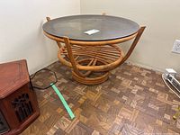 Front side view of the round rattan table showing glass top, rattan base, and rattan slatted shelf beneath.