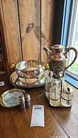 Full set of silverplate items displayed on wood table by window: coffee pot, round mirror tray, round serving dish, condiment holder with glass containers, salt and pepper shakers