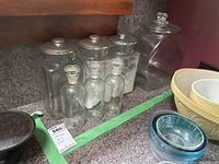 Six vintage glass jars on countertop; three large storage jars with lids and three apothecary bottles with stoppers in front. Glass is clear and transparent.